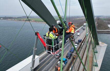 Vier Arbeiter mit Schutzausrüstung auf einer Hebebühne arbeiten an den Tragseilen der Fehmarnsundbrücke.
