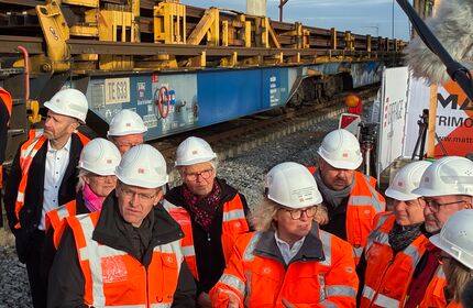 Ministerpräsident Günther, die Projektleitung und andere Stakeholder stehen mit orangenen Westen und weißem Helm vor dem gelben Bauzug