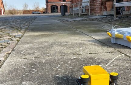 Ein gelber Messsensor steht vor einem Backsteinhaus
