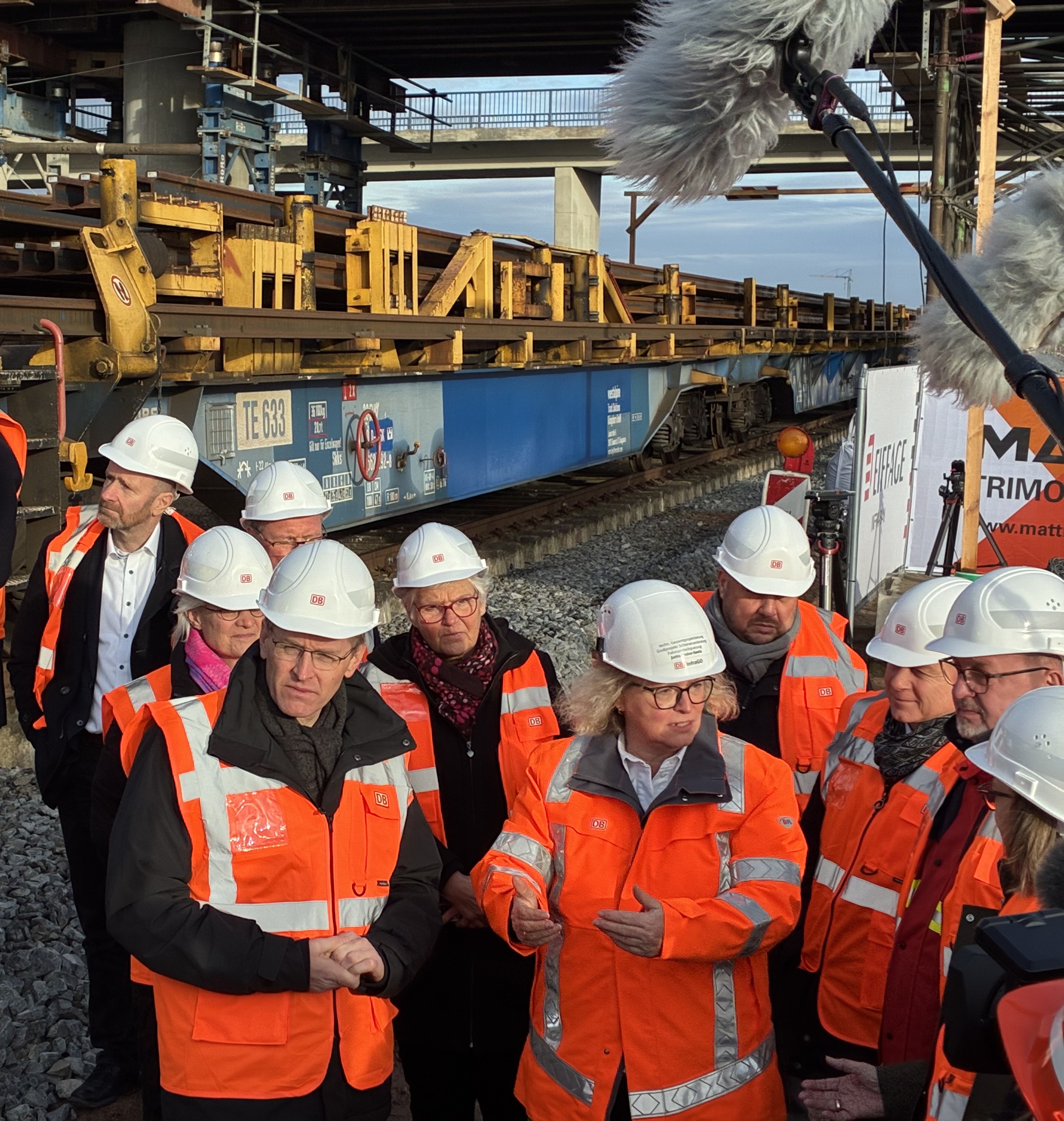 Ministerpräsident Günther, die Projektleitung und andere Stakeholder stehen mit orangenen Westen und weißem Helm vor dem gelben Bauzug