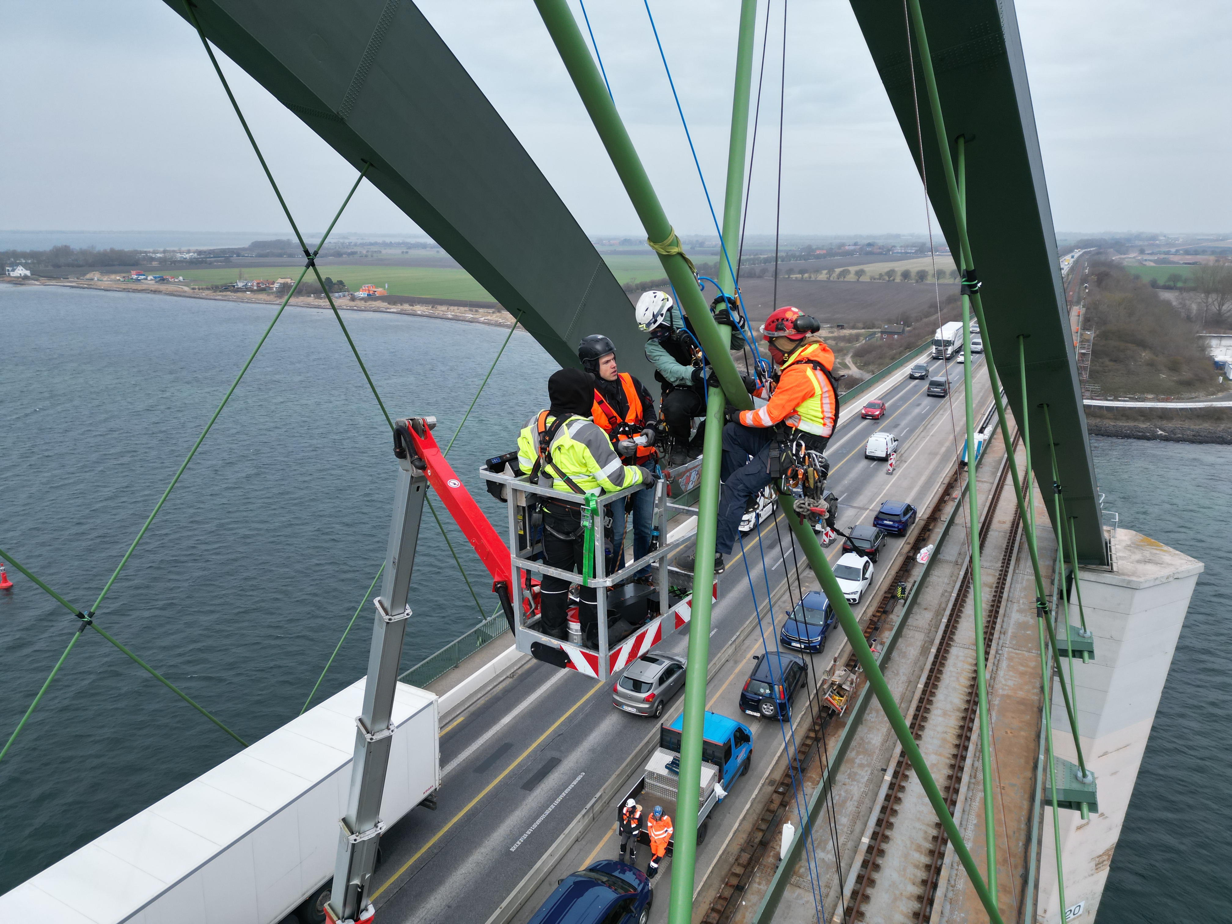 Vier Arbeiter mit Schutzausrüstung auf einer Hebebühne arbeiten an den Tragseilen der Fehmarnsundbrücke.