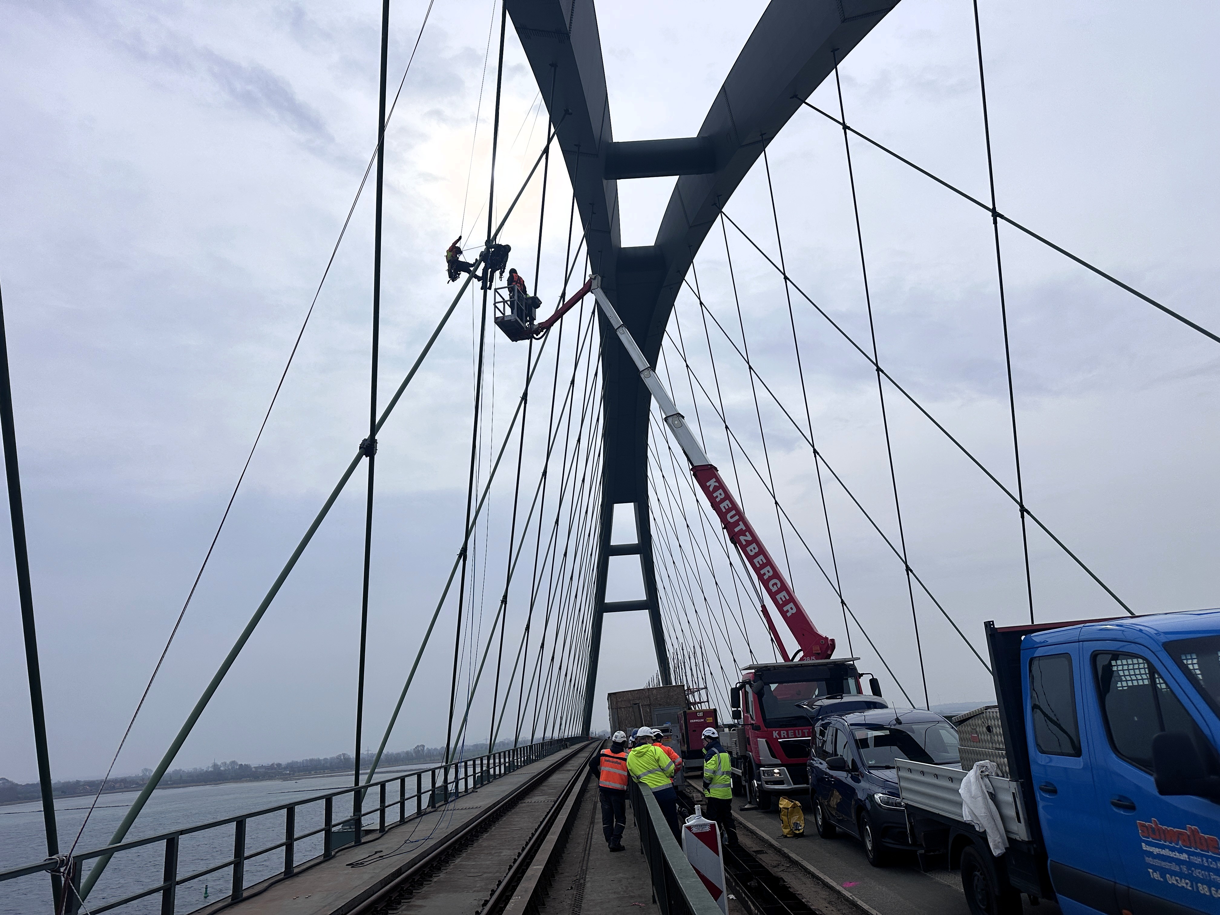 Blick von unten auf die Tragseile der Fehmarnsundbrücke und Arbeiter auf einer Hebebühne.