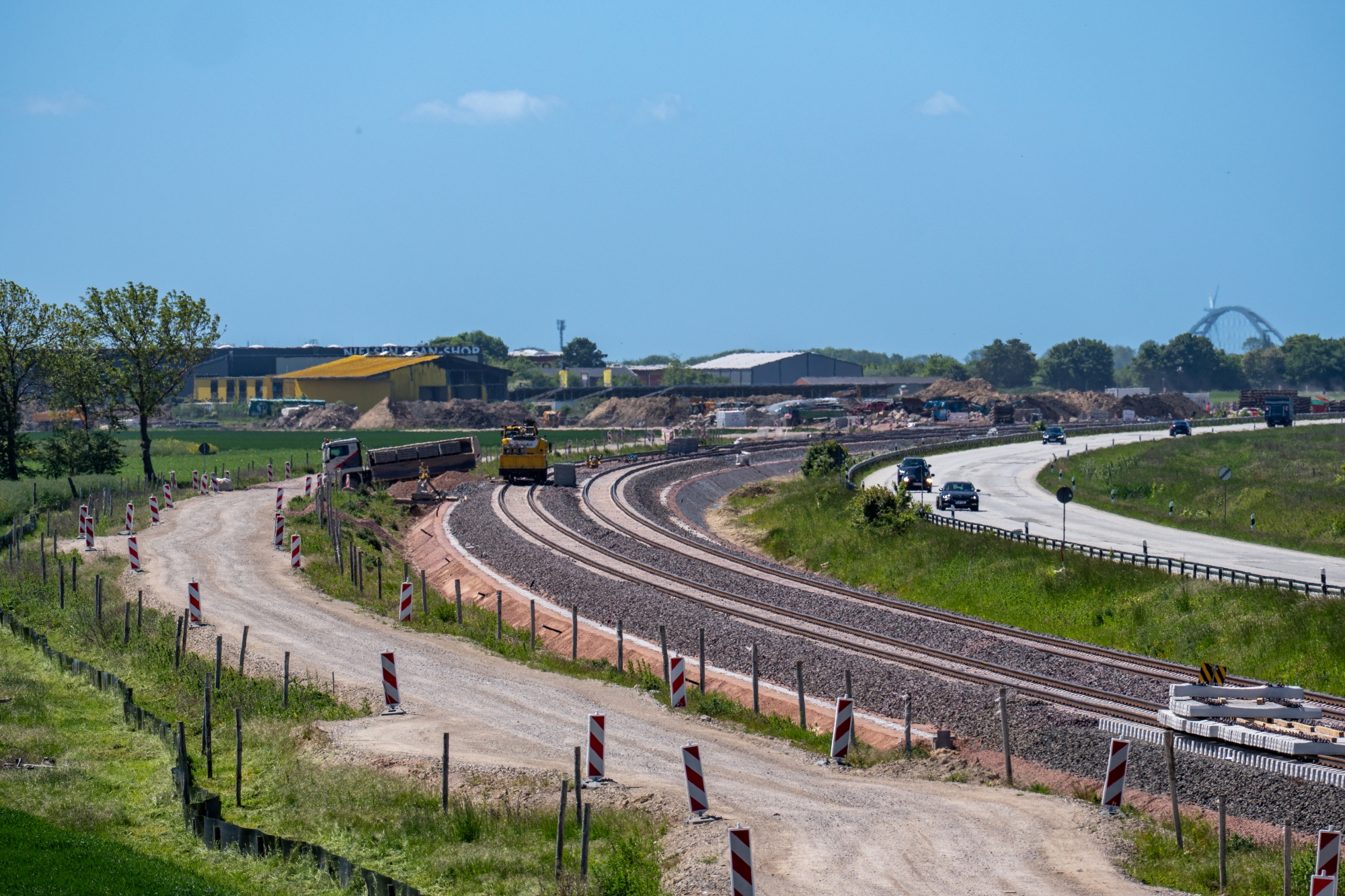 Eine Baustraße und dahinter Gleise auf Schotter auf der Insel Fehmarn, im Hintergrund Häuser, die Bundesstraße und die Fehmarnsundbrücke