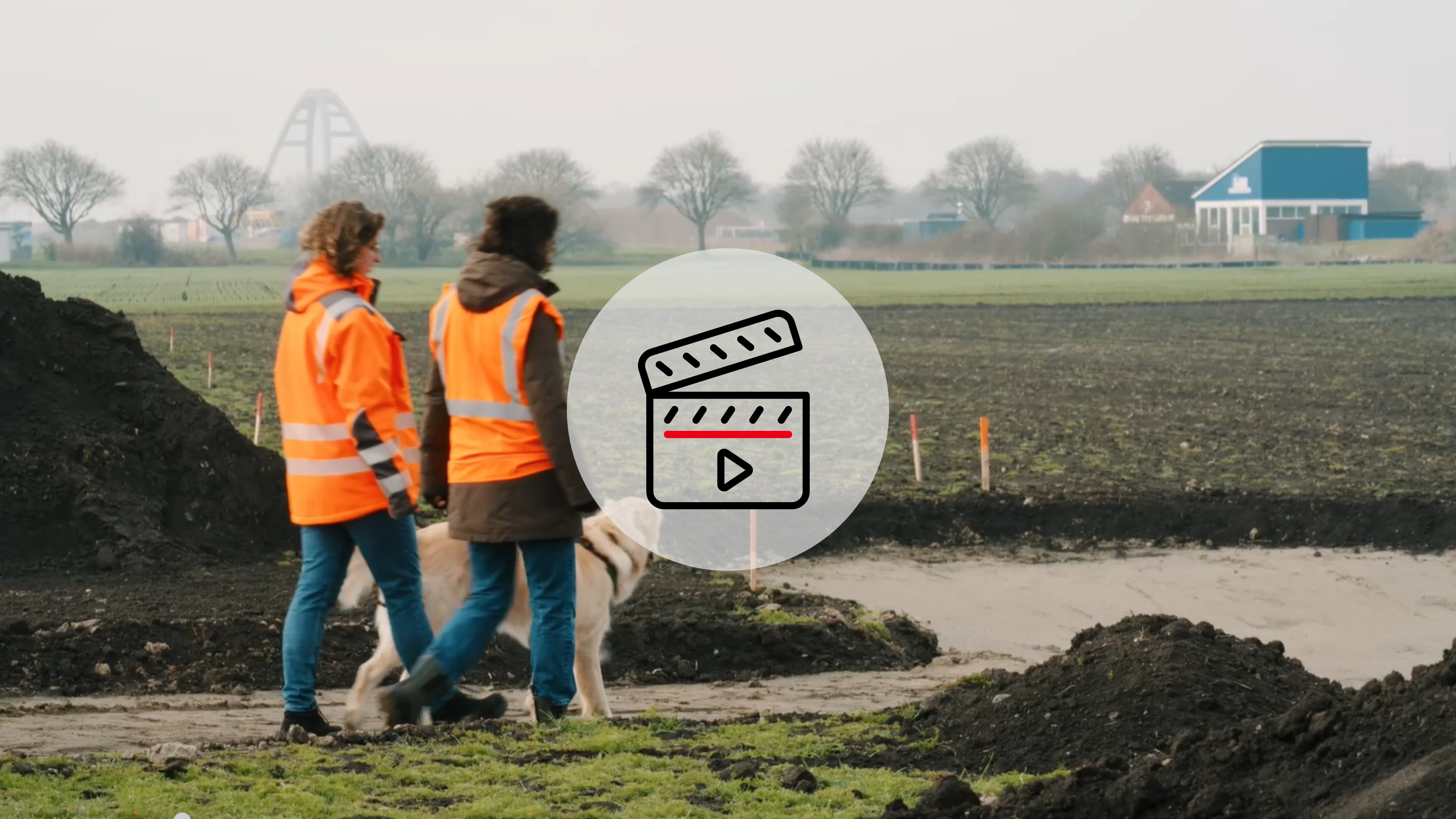 Im Vordergrund Video-Icon, im Hintergrund gehen zwei Frauen mit orangenen Warnwesten und einem hellbraunen Hund über ein Acker auf Fehmarn