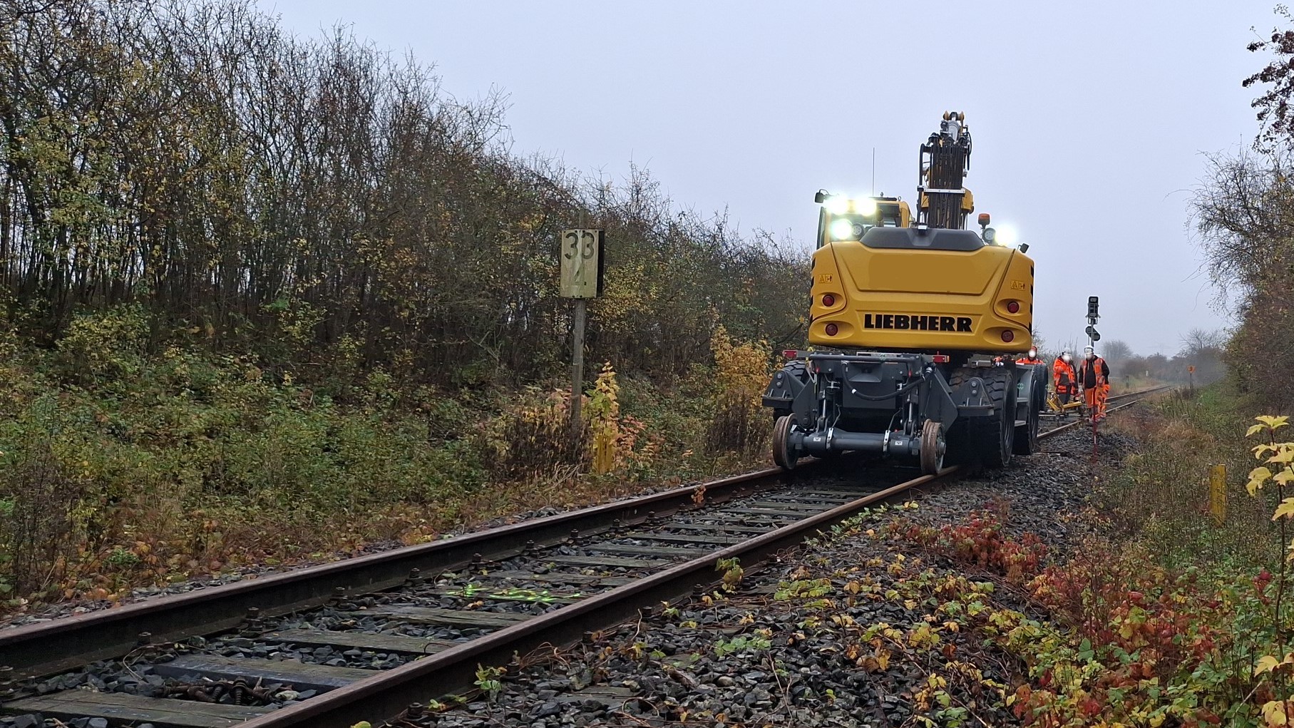 Die Rückseite eines Zweiwegeschienenfahrzeugs der Firma Liebherr steht auf den Gleisen der Strecke 1100 zusammen mit 4 Gleisarbeitern