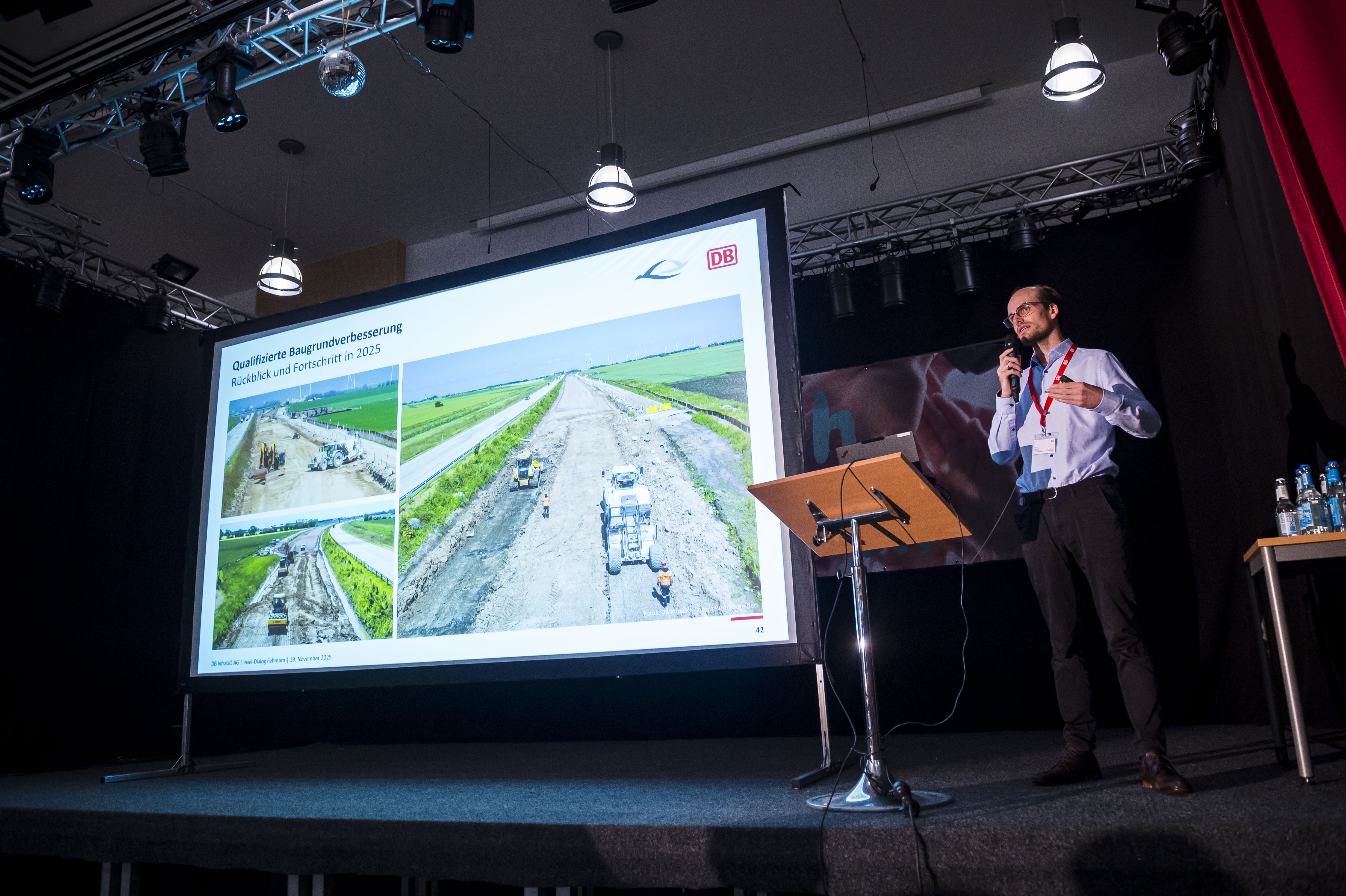 Ein Mann steht auf einer Bühne vor einem Rednerpult und neben einer Leinwand, auf der eine Präsentation zu sehen ist.